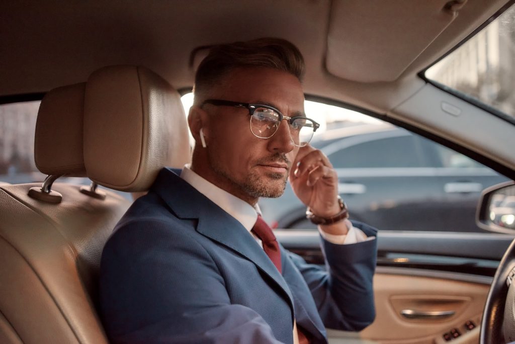 Man in a tailored suit sitting in a luxury car, wearing glasses and AirPods, exuding confidence and sophistication, reflecting the upscale male plastic surgery services offered in New York City.